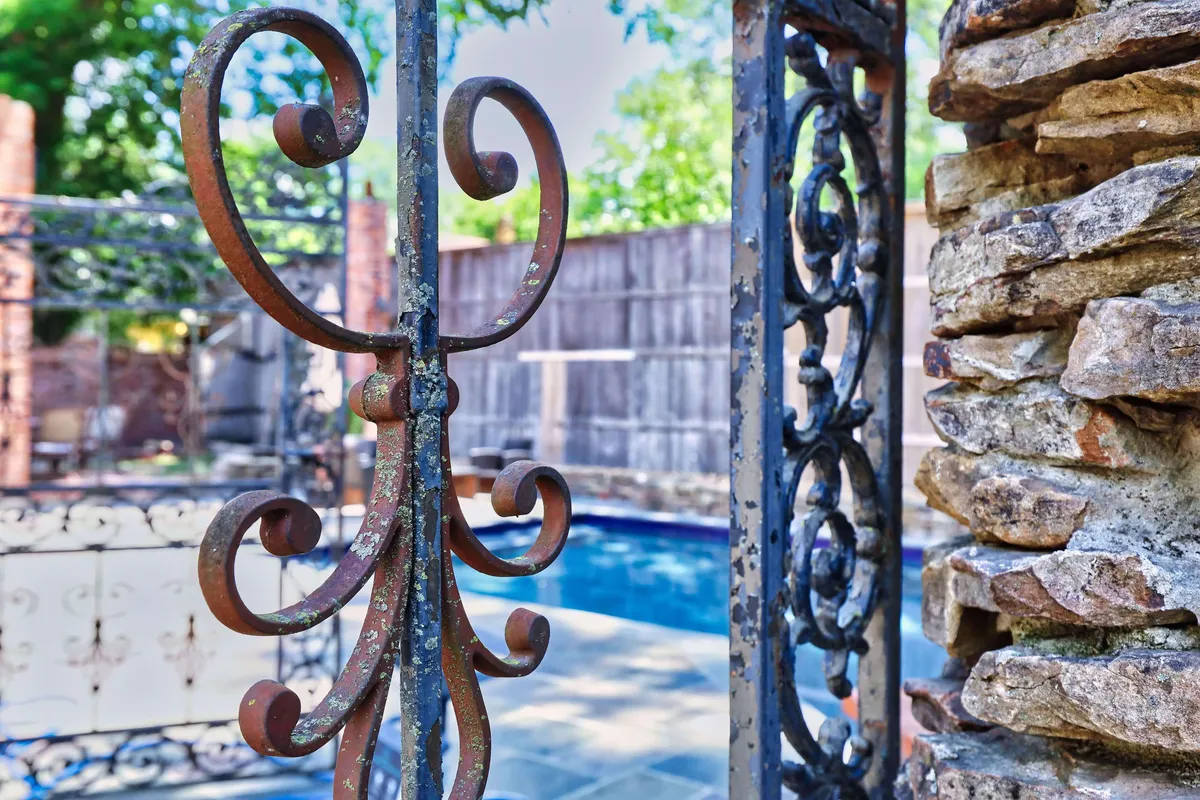 A stylish pool area viewed through an ornate iron gate, with textured stonework and glimpses of the sparkling swimming pool beyond. The scene suggests a tranquil yet secure outdoor space ideal for relaxation or entertaining.
