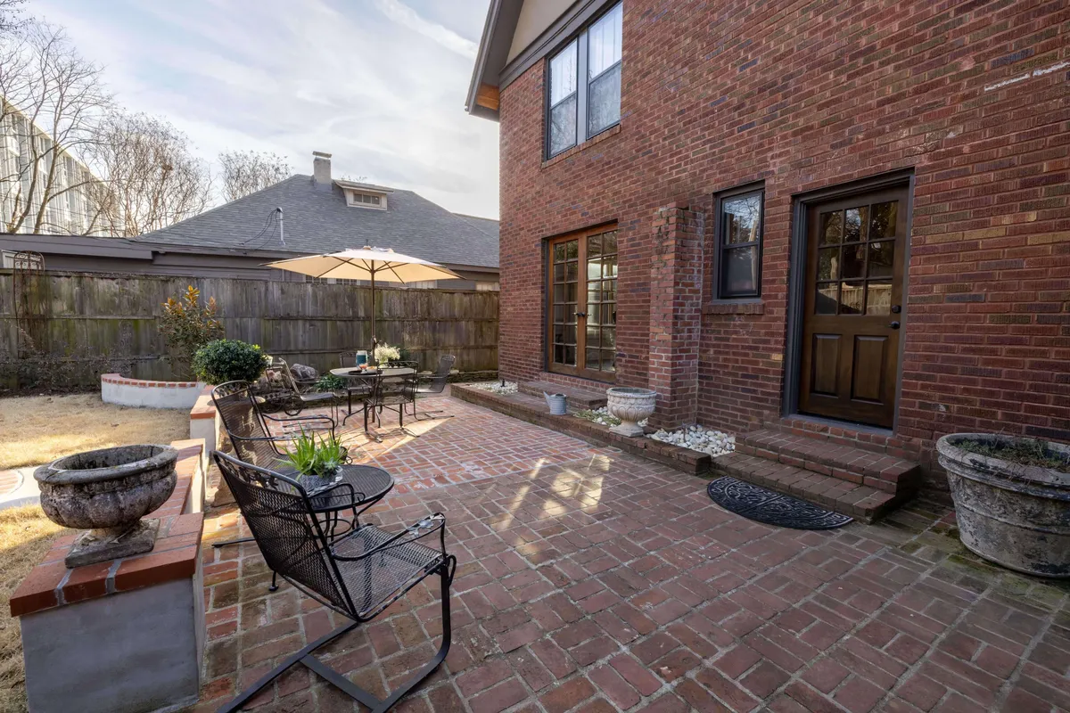 A brick patio with comfortable outdoor seating and a large umbrella sits surrounded by flowering pots and greenery. The space is adjacent to the house, creating a seamless transition from indoor to outdoor living. The setting is ideal for dining, relaxing, or entertaining guests in a peaceful, garden-filled environment.