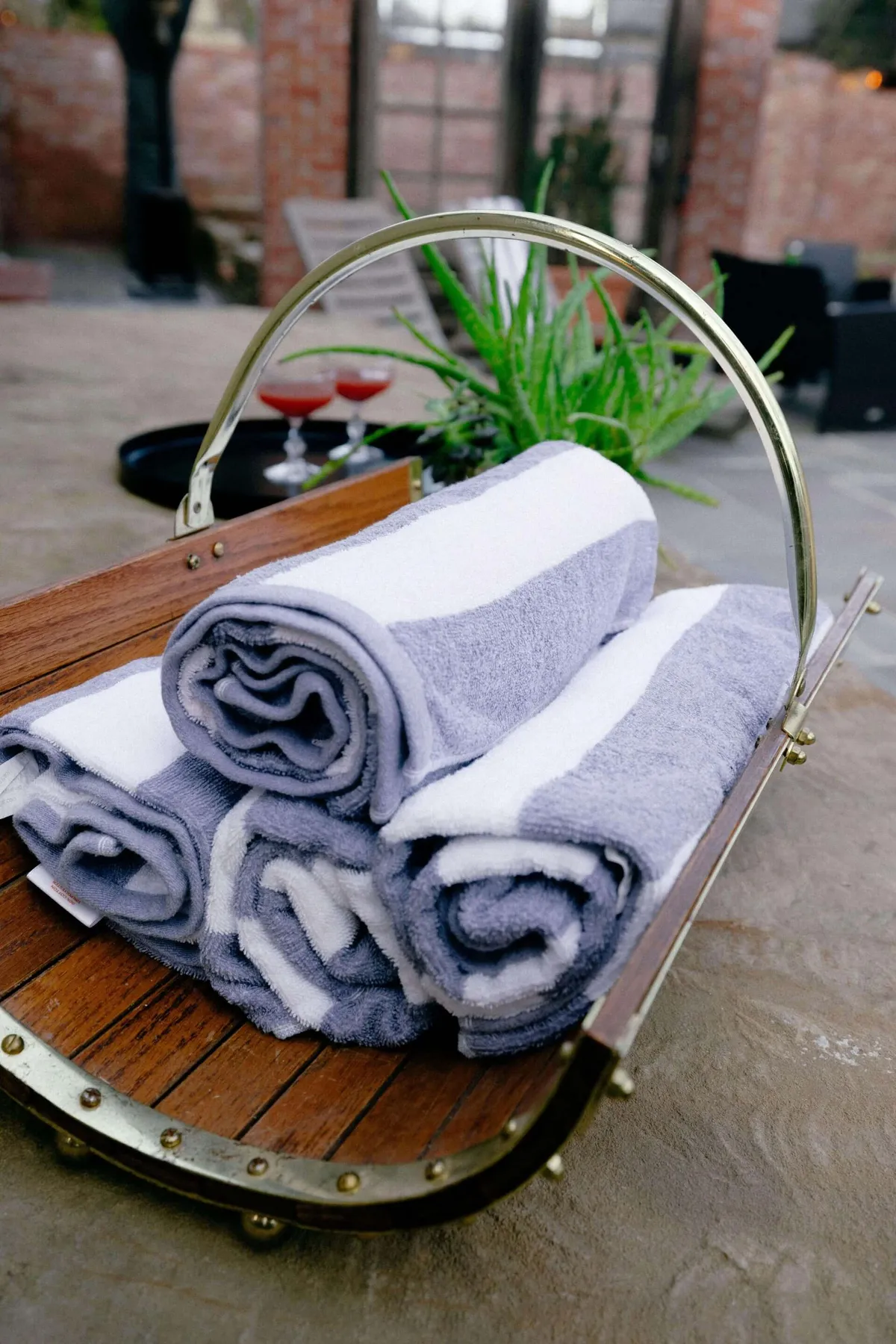 A serene space with neatly stacked towels in a wooden basket, a green plant, and cocktails set out—suggesting a poolside or spa-like atmosphere. The scene is bright, inviting, and evokes leisure and comfort.