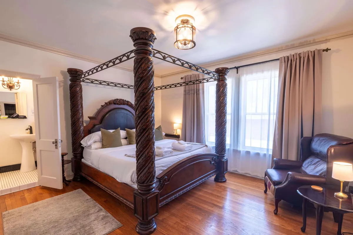 A bright and restful bedroom with a four-poster bed, a comfortable armchair beside a window draped with curtains, and ambient decorative lighting. The space feels both classic and inviting, with soft natural light enhancing the calming atmosphere.