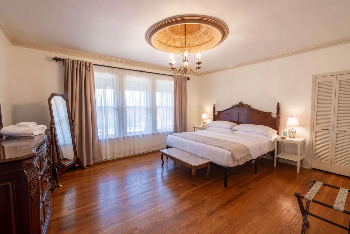 A serene bedroom showcasing a plush bed dressed in neutral linens, a classic chandelier overhead, a large mirror, and a traditional dresser. Natural light filters in, highlighting the elegant and peaceful ambiance, perfect for unwinding or enjoying a quiet morning.