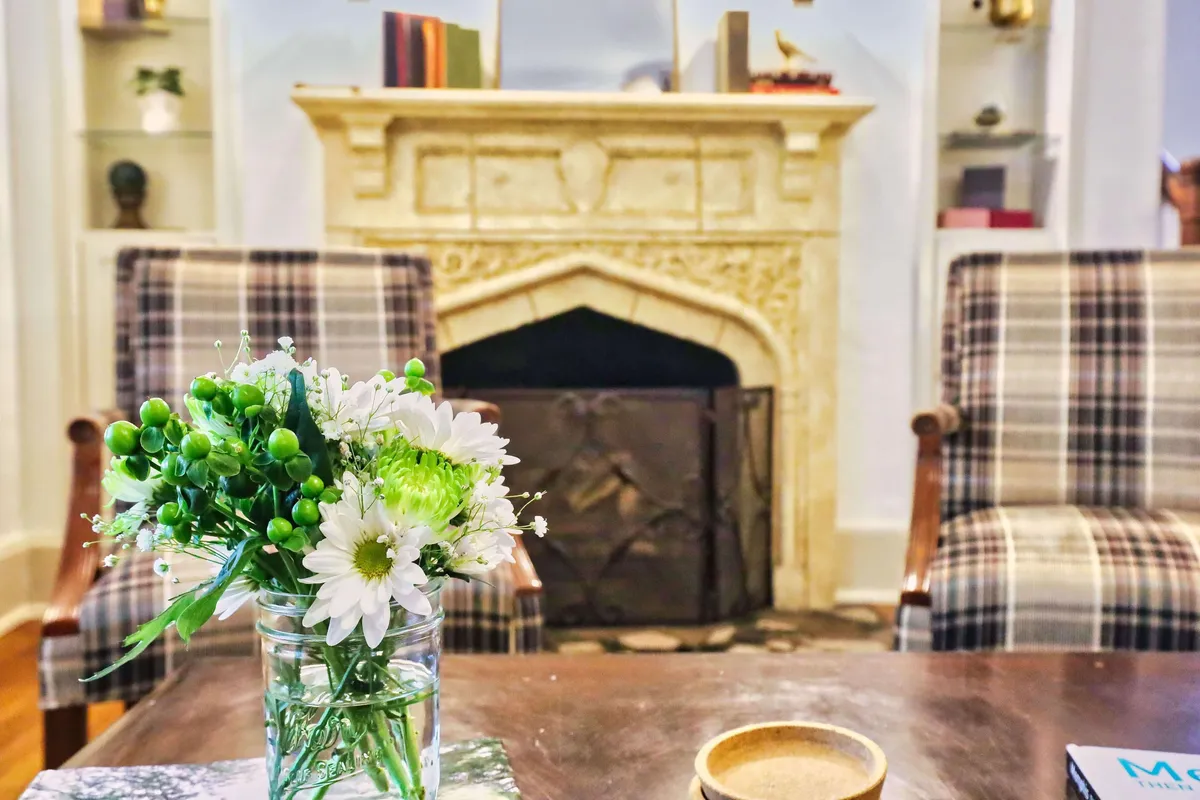 This image showcases a cozy living room with a flower arrangement on the table, a fireplace as the focal point, and plaid armchairs arranged for conversation. The atmosphere is intimate and welcoming, highlighting traditional design elements and warm lighting.