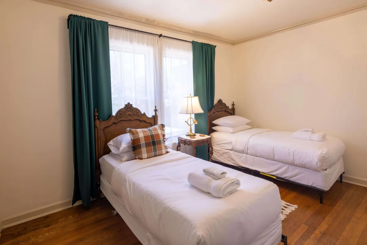A cozy, homely bedroom featuring a bed with a decorative headboard, a plaid-accented pillow, side table lamp, and soft curtains. Sunlight gently streams across the space, creating an inviting, lived-in feel.