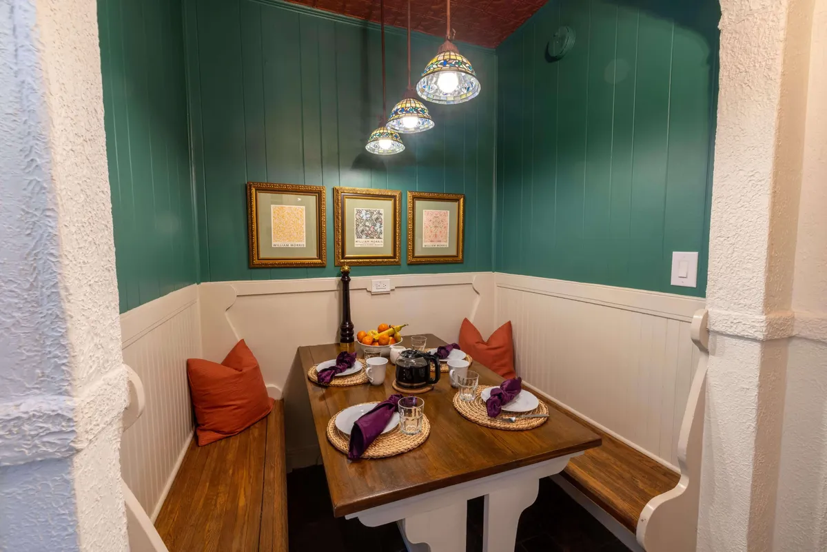 A warm and inviting dining area featuring a wooden table, colorful pendant lights, plush cushions, and artwork. The space is set up for intimate meals, morning coffees, and lively family gatherings.