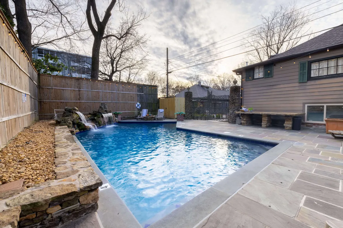 A serene outdoor pool scene featuring a sparkling swimming pool with a waterfall feature, surrounded by lush trees and a stylish seating area. The tranquil setting is enhanced with soft lighting and hints of surrounding nature, inviting relaxation and gatherings.