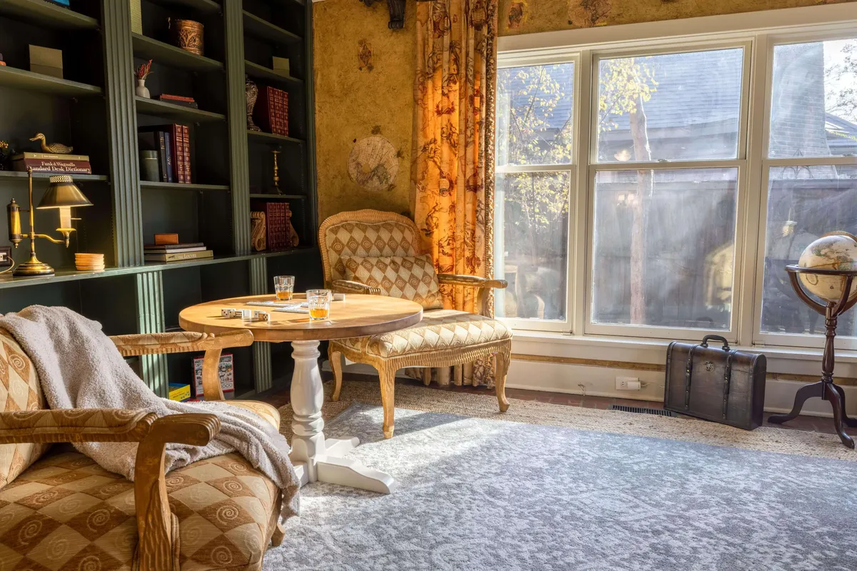 A cozy sitting area with armchairs flanking a coffee table, a bookshelf filled with books, a classic globe, and a large window flooding the room with natural light. The space feels inviting, perfect for relaxed gatherings or quiet moments.