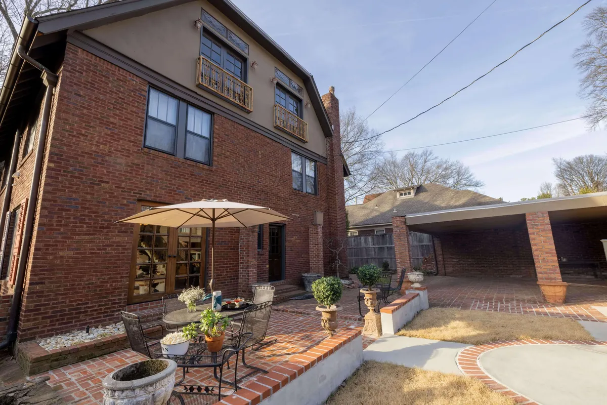 A welcoming exterior view of the estate, featuring the brick facade, outdoor seating area with an umbrella, planters brimming with greenery, and a decorative stone pathway leading toward the entrance. The scene evokes a sense of arrival and outdoor enjoyment.