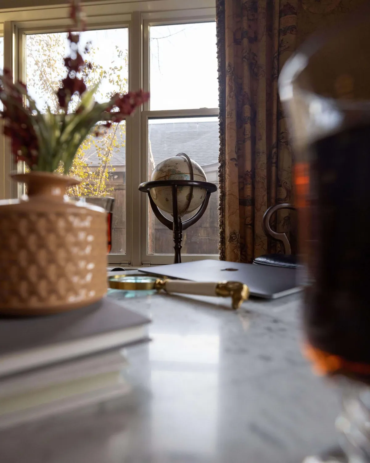 A peaceful workspace set with a marble table, a laptop, a globe, potted flowers, and a magnifying glass, all situated by a window. The mood is reflective and quiet, ideal for working remotely or enjoying a moment of calm.