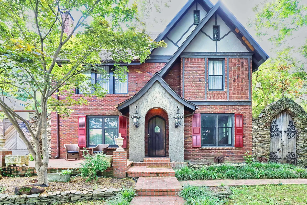 An inviting exterior view of the estate, showcasing an exquisite stone archway, red shutters, and an ornate door nestled within a beautifully landscaped garden. The lush greenery and classic architectural details set a sophisticated and private tone.